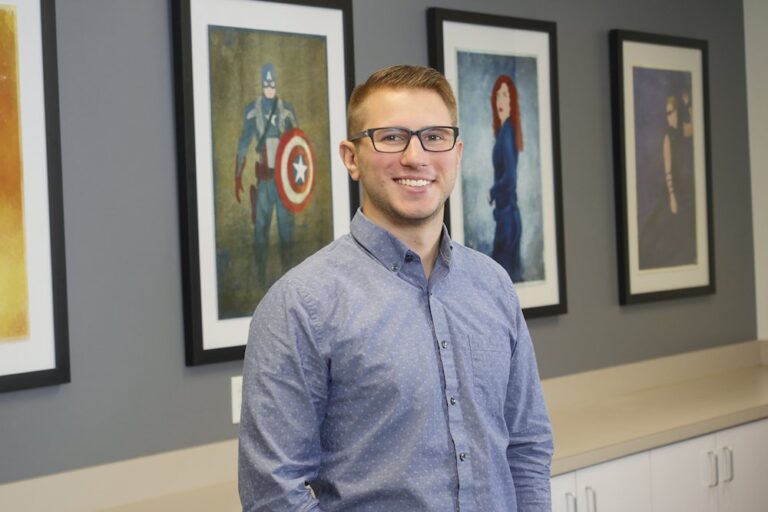 A man wearing glasses and a blue button-up shirt stands in front of a wall with framed superhero artwork.
