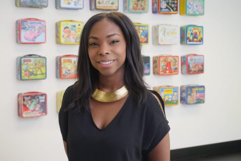 A woman with long dark hair and a gold necklace stands in front of a wall displaying colorful vintage lunchboxes.