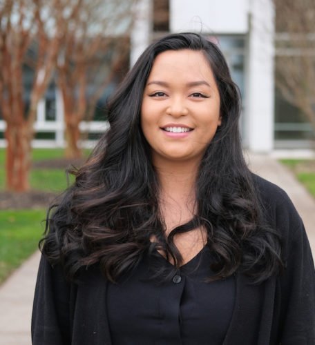 A woman with long, wavy black hair wearing a black top stands outdoors, smiling at the camera. Trees and a building appear in the background.