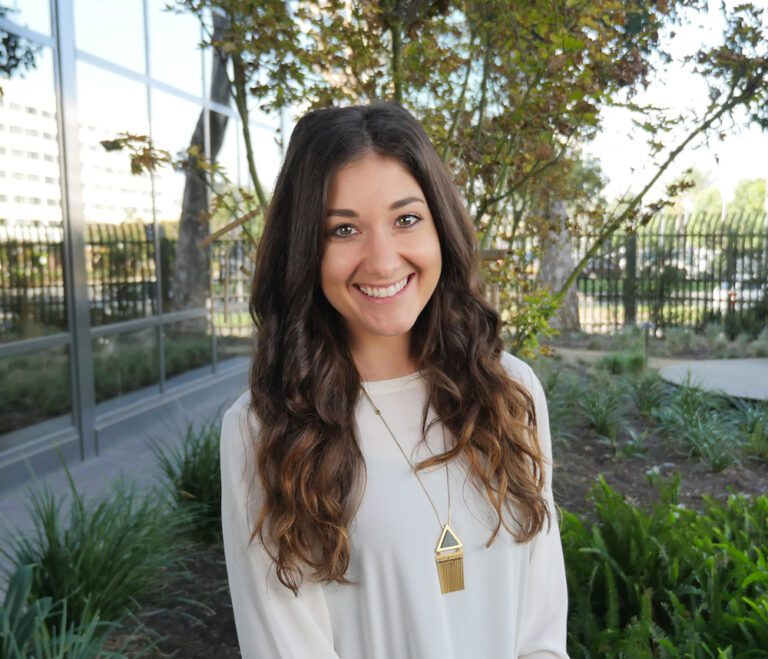 A woman with long brown hair wearing a white blouse and gold necklace stands outside in front of greenery and a glass building, smiling at the camera.