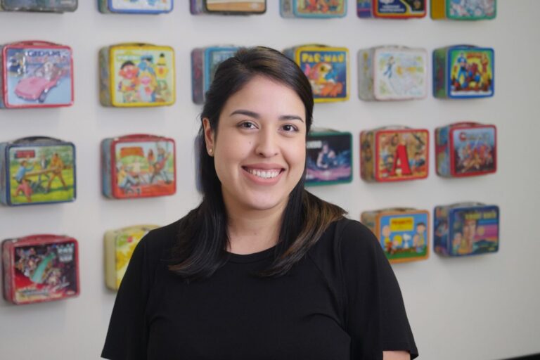 A woman with dark hair smiles at the camera while standing in front of a wall display of colorful vintage lunchboxes.