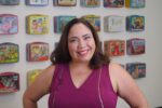 A woman in a magenta sleeveless top smiles in front of a wall decorated with vintage metal lunchboxes.