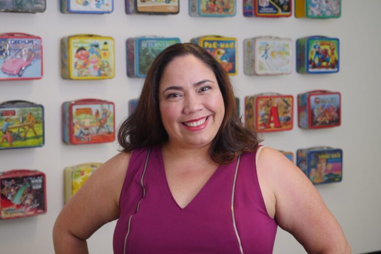 A woman in a magenta sleeveless top smiles in front of a wall decorated with vintage metal lunchboxes.