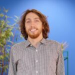 A man with shoulder-length brown hair and a beard, wearing a checkered shirt, stands in front of a blue background with green plants.