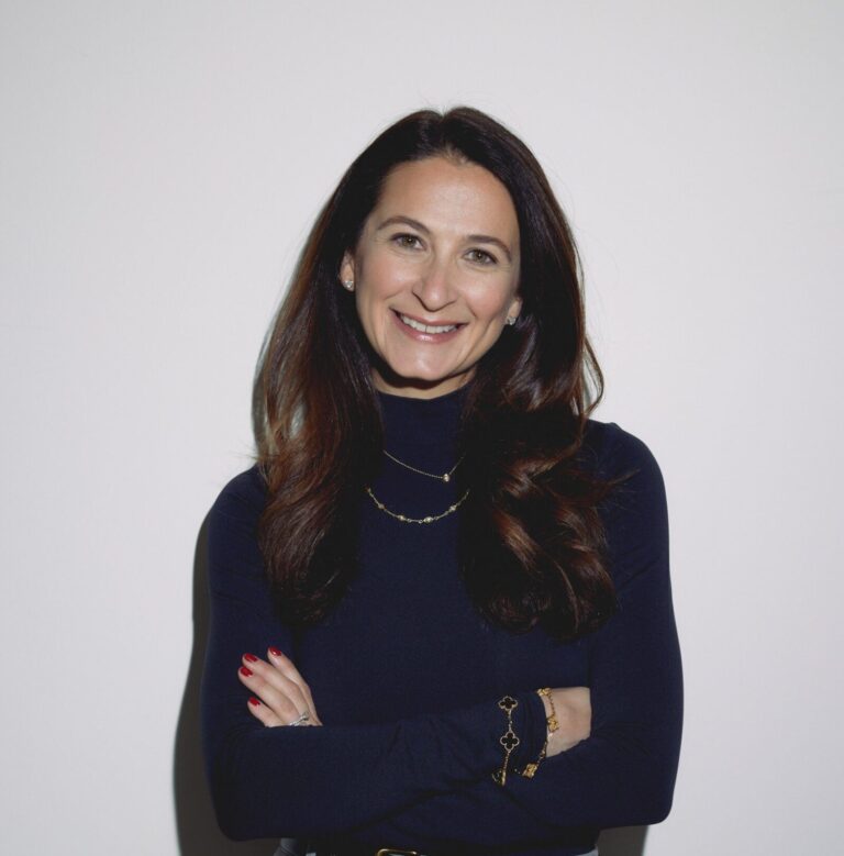 Woman with long brown hair in a black turtleneck top stands against a plain white background, smiling with her arms crossed.