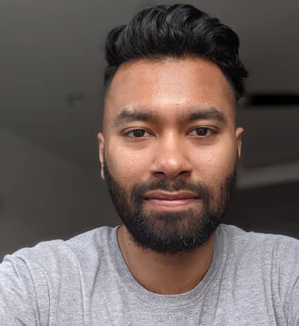 A man with short black hair and a beard is wearing a light grey t-shirt and looking directly at the camera. The background is softly lit and out of focus.