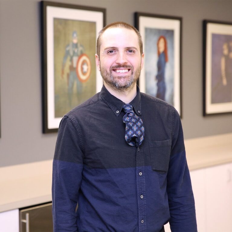 A man with a short beard and dark shirt stands indoors, smiling, with framed superhero artwork visible on the wall behind him.