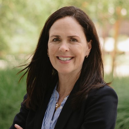 Woman with long dark hair wearing a dark blazer and striped shirt, smiling outdoors with greenery in the background.