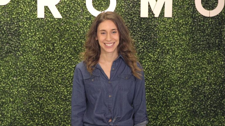 A woman with long, wavy hair wearing a blue button-up shirt stands and smiles in front of a green leafy background with partial white letters visible above her.