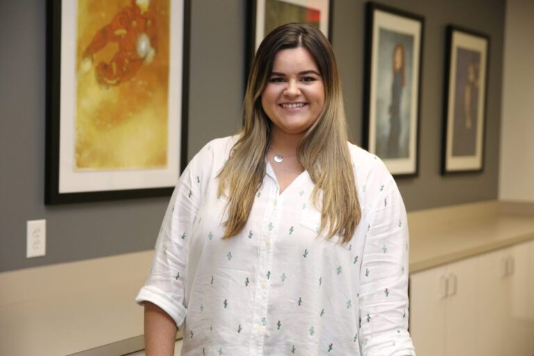 A woman with long brown hair wearing a white shirt stands indoors, smiling, with framed artwork on the wall behind her.