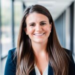 A woman with long brown hair, wearing a dark blazer and white top, smiles while standing in a brightly lit hallway with large windows.