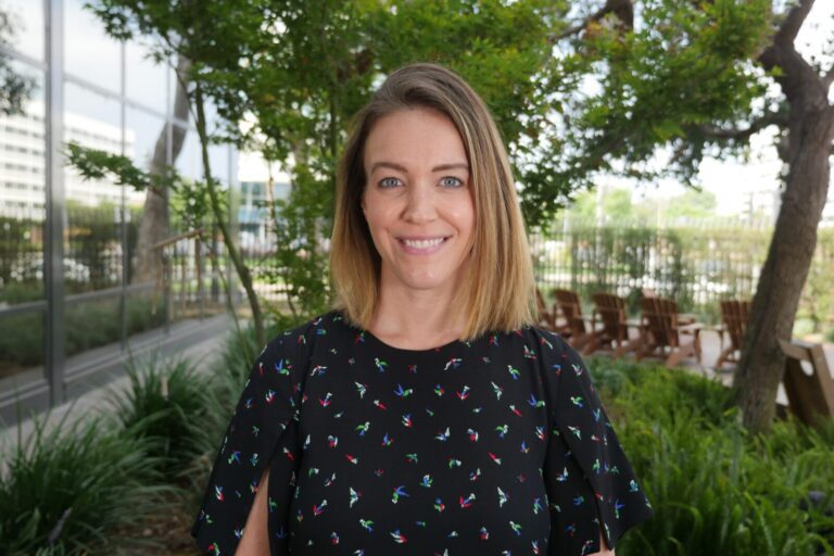 A woman with shoulder-length blonde hair stands outside in front of greenery and patio chairs, smiling at the camera.