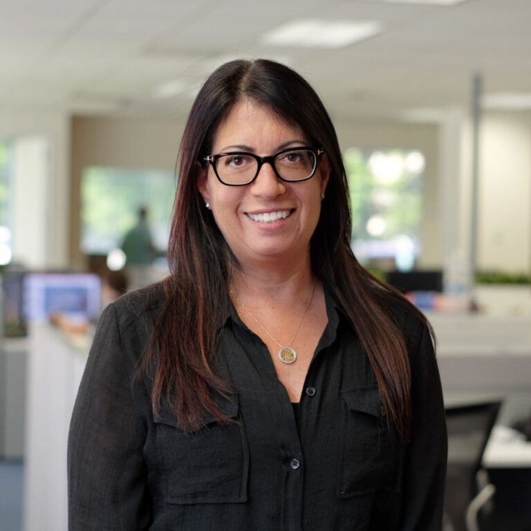 A woman with long dark hair and glasses, wearing a black blouse, stands smiling in a modern office with desks and computers in the background.