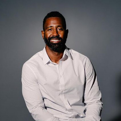 A man with a beard and mustache wearing a white button-up shirt sits and smiles against a plain gray background.