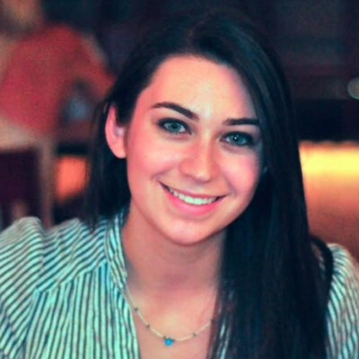 A young woman with long dark hair smiles at the camera, wearing a striped shirt and a necklace, seated indoors with a blurred background.