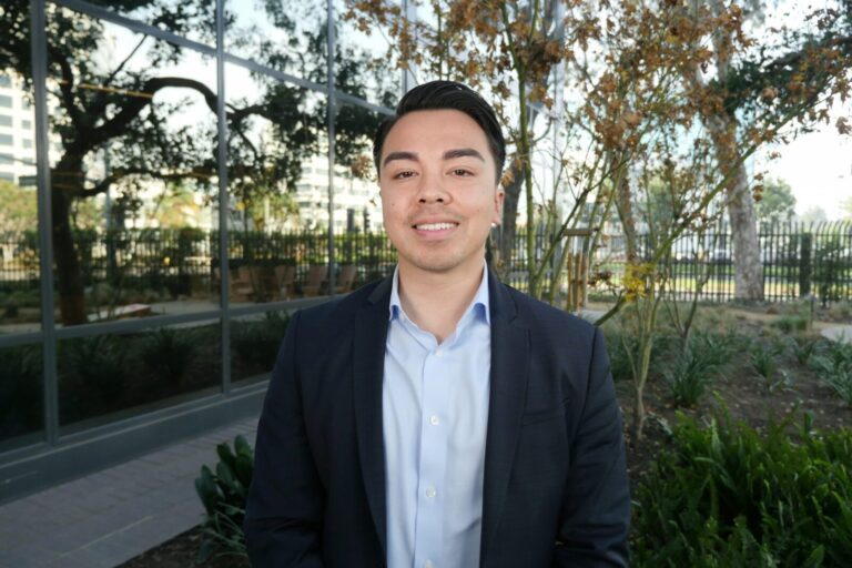 A person in a suit stands outdoors in front of a building with large windows, surrounded by plants and trees.