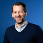 A man with short brown hair and a beard, wearing a light blue shirt and a navy sweater, smiles in front of a plain blue background.