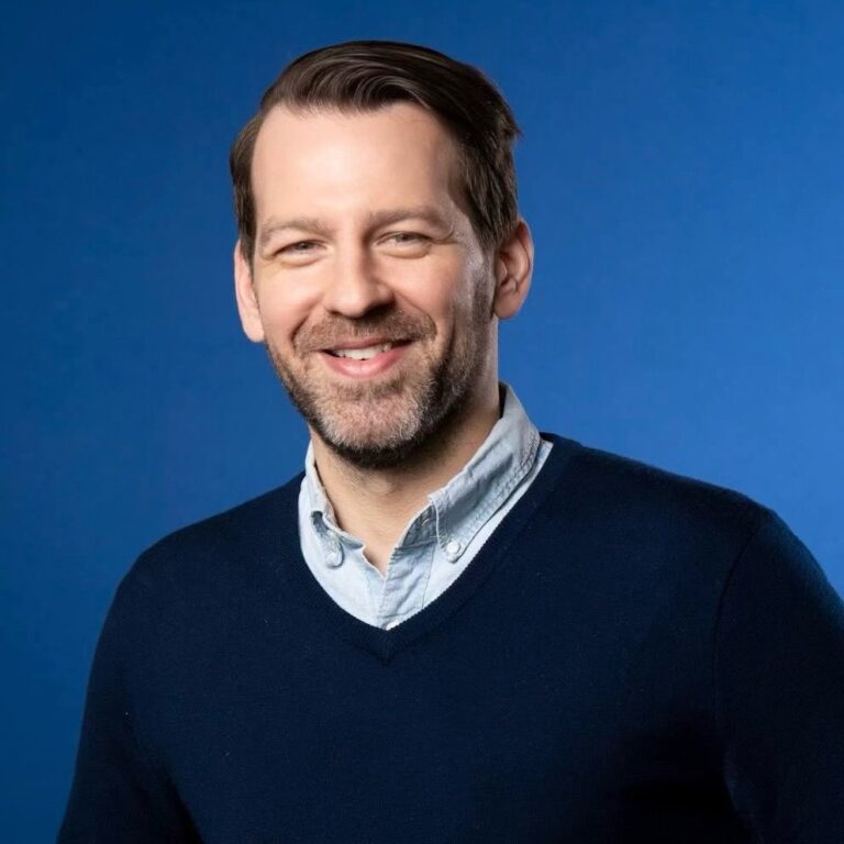 A man with short brown hair and a beard, wearing a light blue shirt and a navy sweater, smiles in front of a plain blue background.