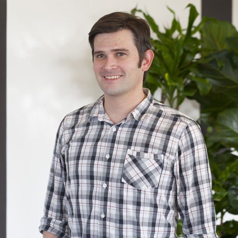 A man with short dark hair, wearing a black and white plaid shirt, stands indoors in front of green plants and a white wall.