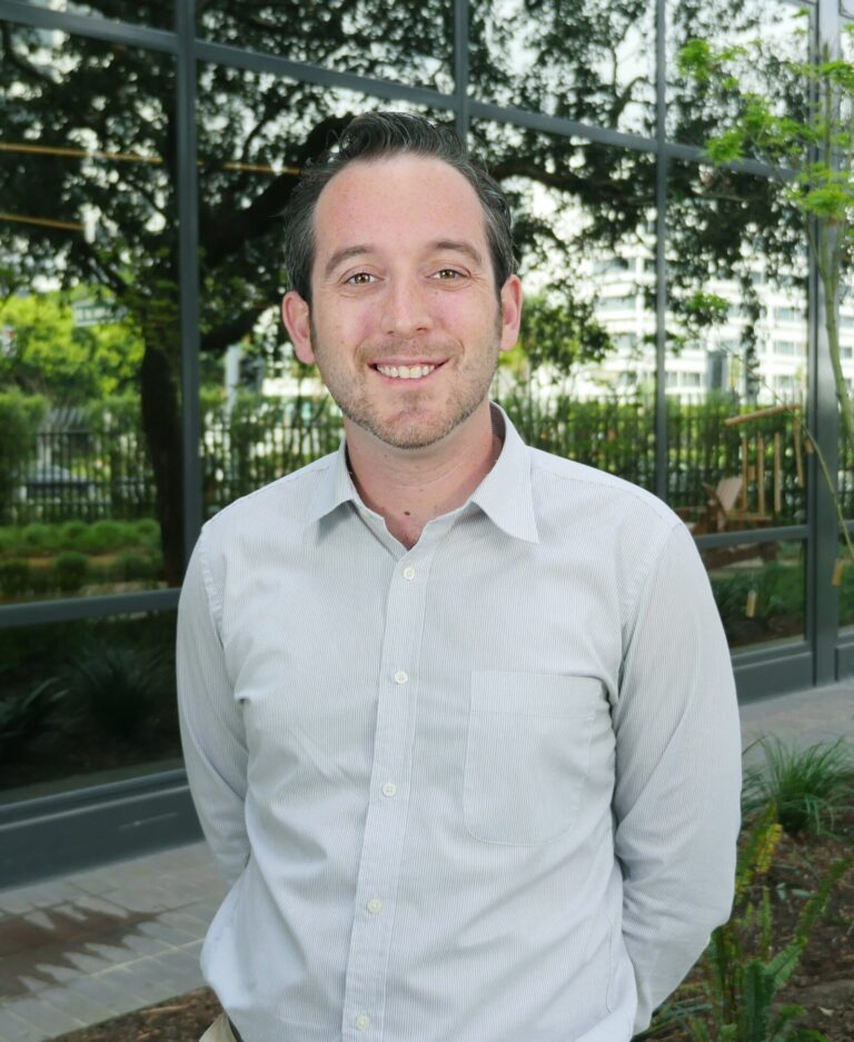A man in a light gray shirt stands outdoors in front of a building with large windows and greenery.