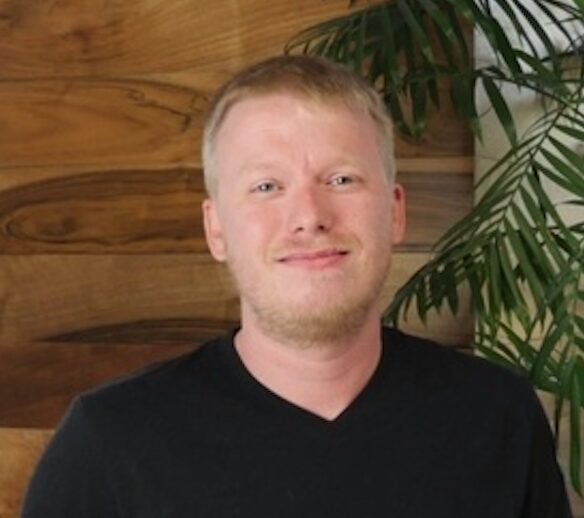 A man with short blond hair and a light beard wearing a black T-shirt stands in front of a wooden wall and a leafy plant.
