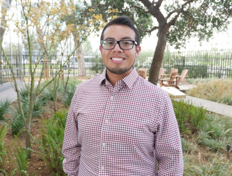 A man wearing glasses and a checked shirt stands outdoors in a garden area with trees, plants, and wooden chairs in the background.