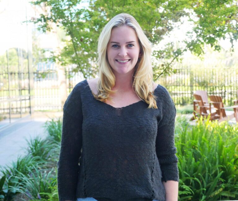 A woman with blonde hair in a dark sweater stands outdoors, smiling, with greenery and wooden chairs in the background.