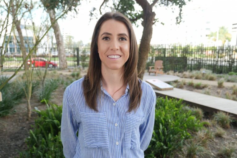 A woman with long brown hair wearing a blue and white striped shirt stands outdoors in a garden area, smiling at the camera.