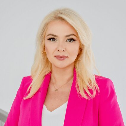 A woman with long blonde hair wearing a bright pink blazer and white top stands against a plain light background, looking directly at the camera.