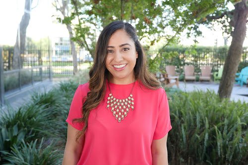 A woman with long brown hair, wearing a pink top and a statement necklace, smiles while standing outdoors in a garden with trees and chairs in the background.