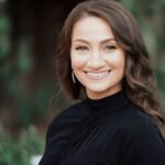 A woman with long wavy brown hair, wearing a black top and earrings, smiles at the camera with a blurred outdoor background.