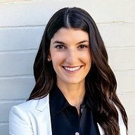 A woman with long dark hair, wearing a white blazer over a black shirt, stands in front of a light-colored brick wall, smiling at the camera.