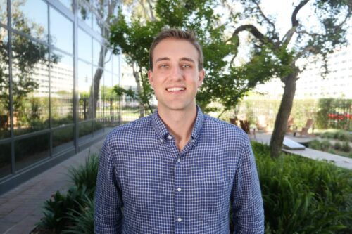 A man wearing a blue checkered shirt stands outdoors in front of greenery and a glass building, smiling at the camera.
