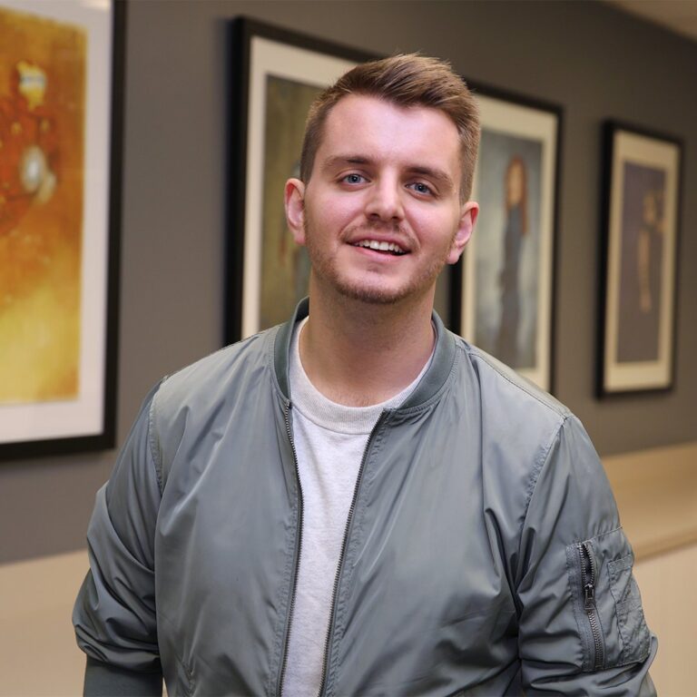 A young man with short hair and a trimmed beard, wearing a light gray bomber jacket over a white shirt, stands indoors in front of framed artwork.