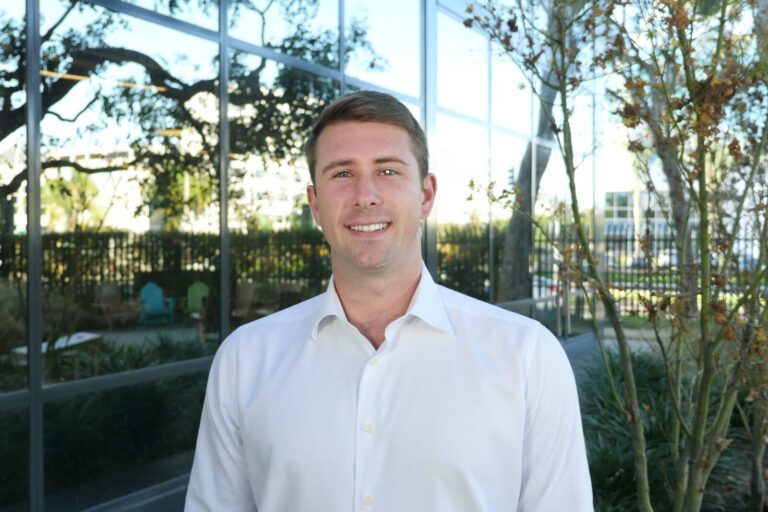 A man in a white shirt stands outside in front of a building with large windows and surrounding greenery.