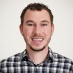 A man with short curly brown hair and a trimmed beard, wearing a black and white plaid shirt, smiles in front of a plain light background.