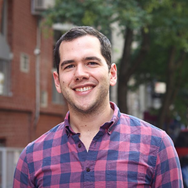 A man wearing a pink and blue plaid shirt stands outside, smiling, with trees and brick buildings in the background.