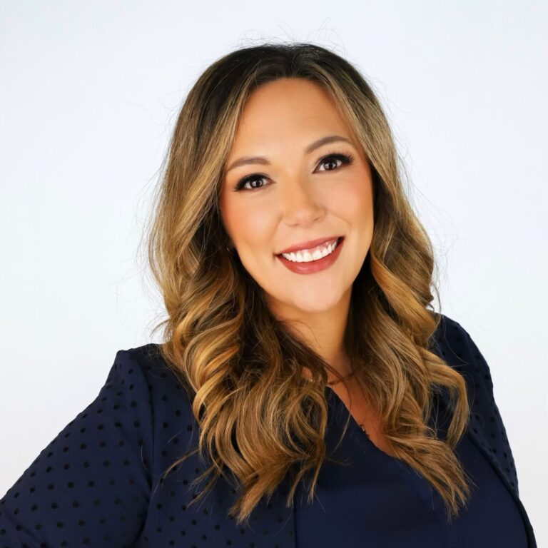 Woman with long wavy blonde hair, wearing a navy blue blouse, smiling and posing in front of a plain white background.