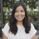 A woman with long dark hair and a white blouse smiles while standing outdoors in front of glass windows and greenery.