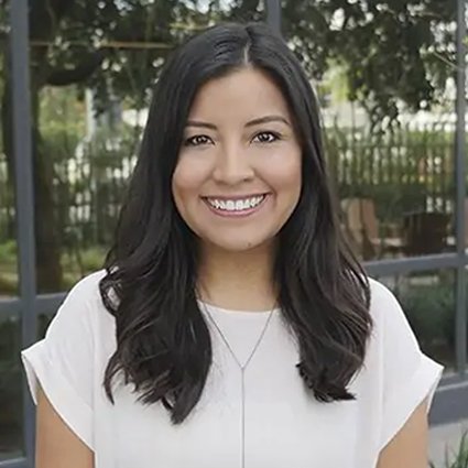 A woman with long dark hair and a white blouse smiles while standing outdoors in front of glass windows and greenery.