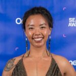 A woman with long earrings and a gold halter dress smiles in front of a blue step-and-repeat event backdrop.