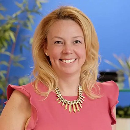 A woman with blonde hair wearing a pink top and a statement necklace smiles in front of a blue background with plants.