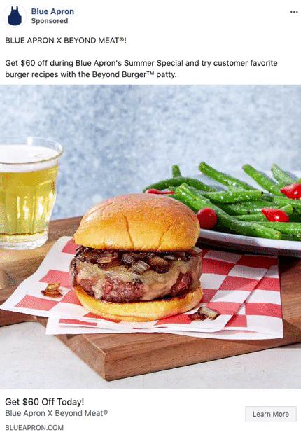 A Beyond Burger patty sandwich with grilled onions is served on a wooden board with a side of green beans and tomatoes, alongside a glass of light beer.