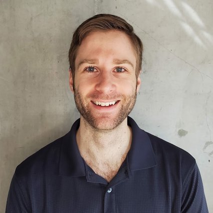 A man with short light brown hair and a beard smiles at the camera, standing against a plain, light-colored concrete wall.