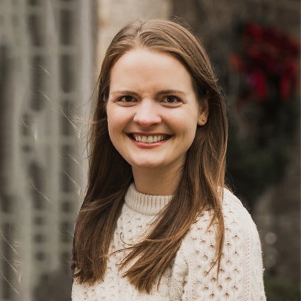 A woman with straight brown hair wearing a white knit sweater stands outdoors, smiling at the camera.