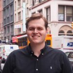 A man wearing a black button-up shirt stands smiling on a city street with buildings, cars, and a yellow bus in the background.