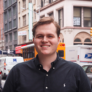 A man wearing a black button-up shirt stands smiling on a city street with buildings, cars, and a yellow bus in the background.