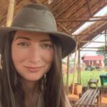 A woman wearing a wide-brimmed hat sits under a thatched roof structure, with greenery and a wooden house visible in the background.