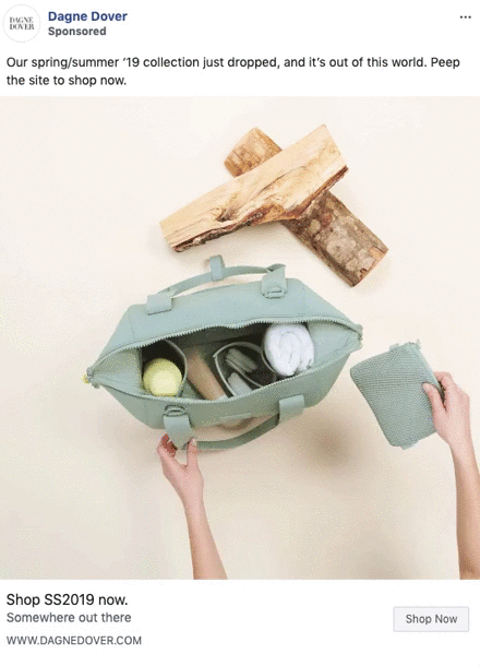 A person places a small zippered pouch into a light green handbag, which contains a towel, bottle, and other items. Two logs are arranged above the bag on a beige surface.