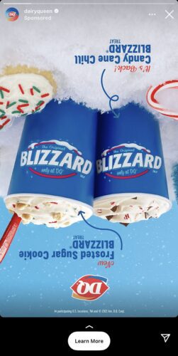 Two Dairy Queen Blizzard cups are shown upside down on a snowy surface beside two spoons and frosted sugar cookies with sprinkles.